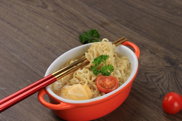 Close-up Indomie, smakelijke Indonesische Instant Noodle met ei, tomaat en gehakte chili op oranje kom met Chopstick. Op de tafel met kopieerruimte