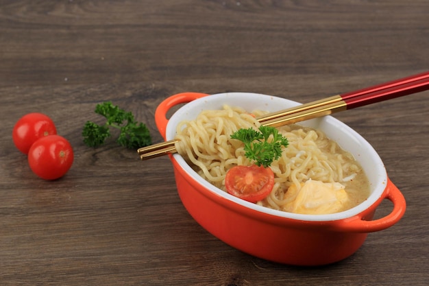 Indomie, 젓가락으로 오렌지 그릇에 계란, 토마토, 다진 고추를 곁들인 맛있는 인도네시아 인스턴트 국수를 닫습니다. 복사 공간이 있는 테이블에서