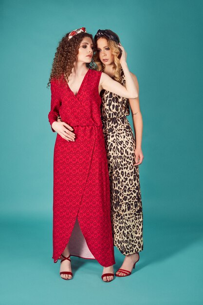 Close up indoor studio fashion portrait of gorgeous women in stylish red and leopard dresses. Blonde and brunette girls isolated on turquoise