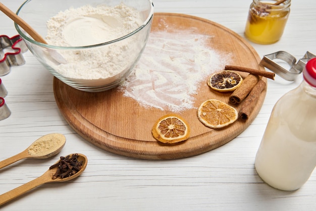 Close-up of ingredients for preparing dough and cooking gingerbread cookies, cutting molds, bottle with plant based milk, glass jar with honey and a bowl with flour on a round wooden board