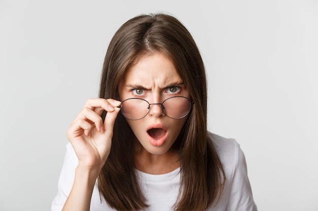 Close-up of insulted and shocked young angry woman looking from under glasses.