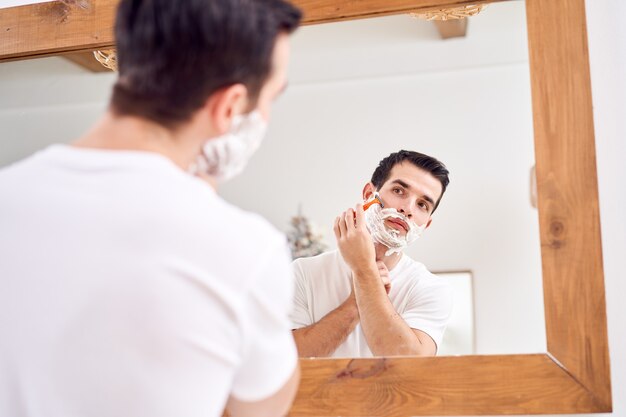 Close-up jonge man in wit T-shirt scheert terwijl hij 's ochtends in de buurt van de spiegel in bad staat