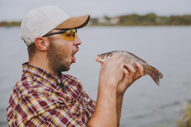 Close-up jonge ongeschoren glimlachende man in geruit hemd, pet en zonnebril ving een vis en opende zijn mond om het te eten aan de oever van het meer op de achtergrond van water. lifestyle, vrijetijdsconcept voor vissers