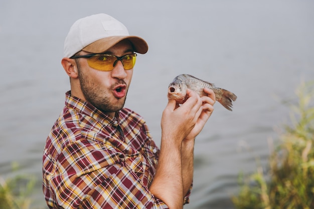 Close-up Jonge ongeschoren man in geruit hemd, pet en zonnebril ving een vis, toont het en verheugt zich aan de oever van het meer op de achtergrond van water. Lifestyle, recreatie, vrijetijdsconcept voor vissers