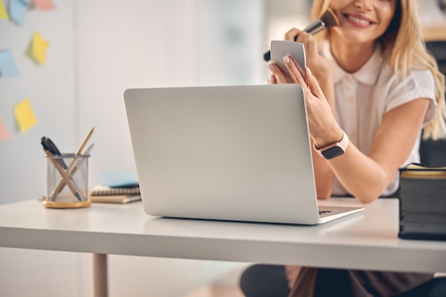 Close up of joyful lady applying face powder with brush while sitting at office table with laptop