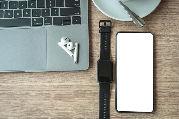 Foto primo piano del computer portatile con tastiera, messa a fuoco degli auricolari wireless con smartwatch, caffè caldo in tazza e schermo vuoto dello smartphone su sfondo in legno scrivania in ufficio nella caffetteria come sfondo