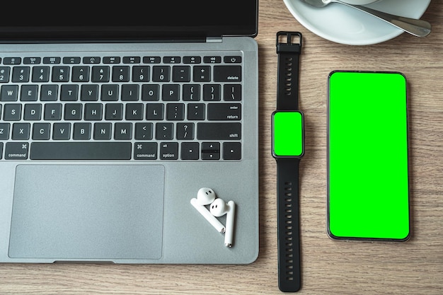 Close-up of Keyboard laptop computer, wireless earphones with smartwatch and smartphone empty screen on wooden background office desk in coffee shop like the background,Green screen