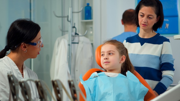 Close up of kid patient with toothache wearing dental bib talking with dentist before intervetion showing to affected mass. Girl sitting on stomatological chair while nurse preparing sterilized tools.