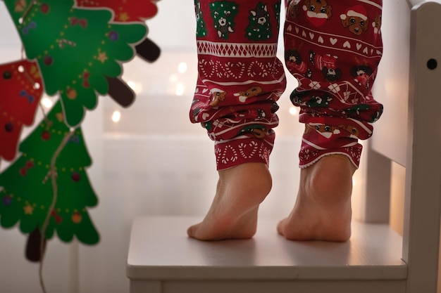 Close-up kind blote voeten. Kid staan op stoel op spitzen versiert raam met lichte kerst slinger en papier kerstboom handgemaakte slinger. Help familie versieren huis