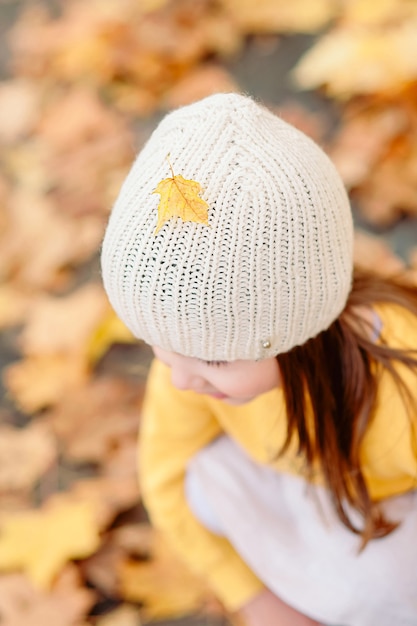 Close-up of a knitted hat with a fallen autumn yellow leaf on a cute girl in the fall