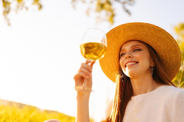 Close-up lage hoekmening van vrolijke gember jonge vrouw in stro hoed met wijnglas op de achtergrond van heldere blauwe hemel op zonnige zomerdag