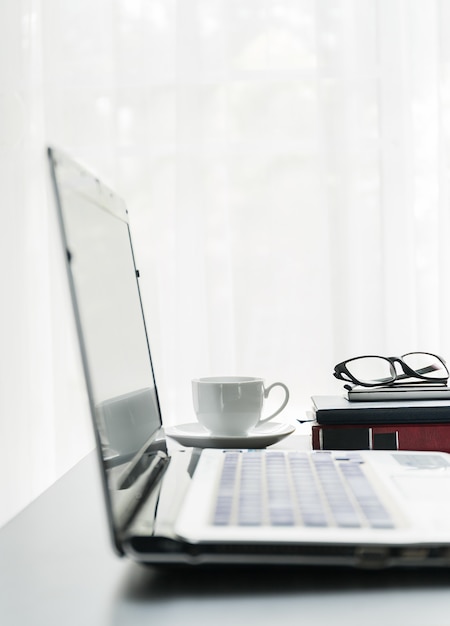 Close up laptop placed on desktop with coffee cup and glasses on book in room at home office