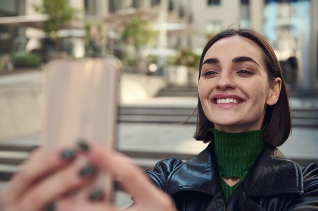 Close-up lifestyle portrait of beautiful funny young brunette with stylish short hair, holding smartphone in outstretched hands and smiling toothy smile taking selfie while standing outdoors in city