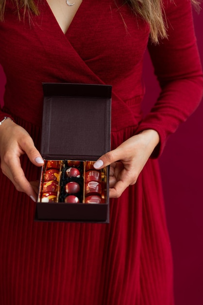 여성의 손에 있는 작은 봉봉 상자를 닫습니다. 작은 상자에 초콜릿 사탕을 복용하는 여성. 레드 스튜디오 배경입니다. 크리스마스 휴가를 위한 달콤한 선물의 개념입니다.
