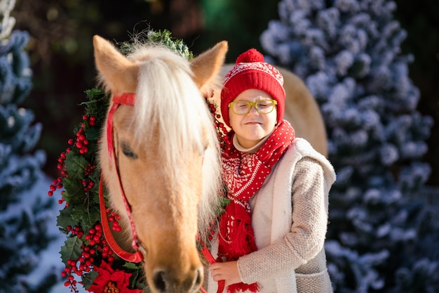 Close-up little boy with glasses and adorable pony with festive wreath near the small wooden house 