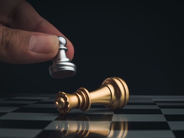 Close-up little silver pawn chess piece wins golden queen and falls on chessboard, dark background. Hand moving chess figure on chess game competition. Strategy, management, and leadership concept.