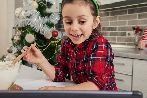 Close-up of a little vlogger, European beautiful baby girl mixing flour in glass bowl, preparing dough, talking by video call, recording video blog against a Christmas tree background at home kitchen