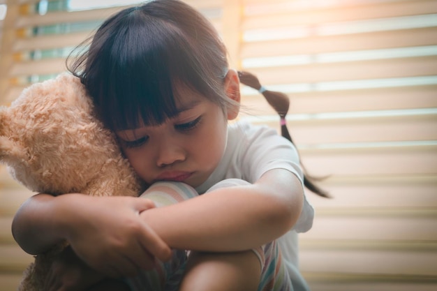 Крупным планом одинокая маленькая девочка обнимает игрушку, сидит дома одна, расстроенный несчастный ребенок ждет родителей, думая о проблемах, плохих отношениях в семье, психологической травме