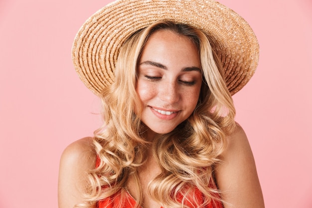Крупным планом симпатичная молодая блондинка в летнем платье и шляпе с ставом позирует изолированной над розовой стеной