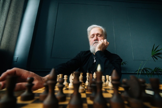 Close-up low-angle view of pensive gray-haired senior aged man thinking game strategy sitting on wooden table with chess board, selective focus. Thoughtful bearded old man playing chess alone at home.