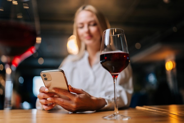 Close-up lowangle weergave van mooie blonde vrouw met behulp van mobiele telefoon typen online bericht zittend aan tafel met glas rode wijn in restaurant