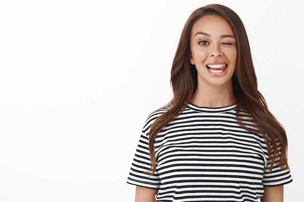 Close-up lucky carefree brunette female in striped t-shirt staying positive, feel accomplished and upbeat, showing tongue playfully, making goofy grimace, standing white wall