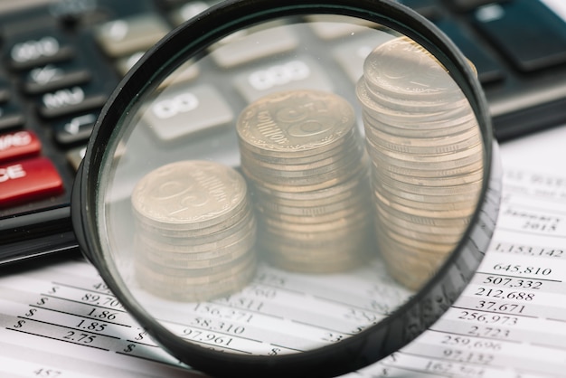 Photo close-up of magnifying glass over the stack coins and calculator on financial balance sheet