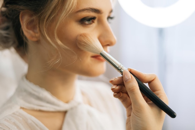 Close-up of make-up artist putting rouge on cheek of young woman using brush while hairdresser making hair-do for young woman in beauty salon. Concept of backstage work.