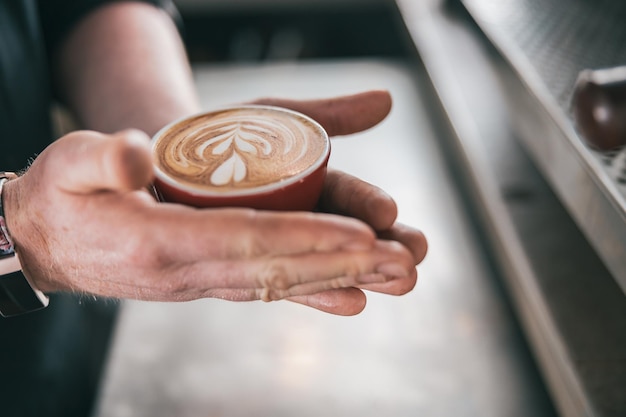 コーヒー ショップで作りたてのカプチーノやラテ カップを保持している男性のバリスタの手のクローズ アップ