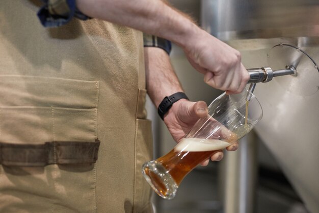 Photo close up of male brewmaster pouring craft beer into glass from tap at brewing factory, copy space