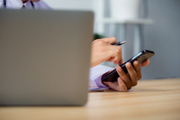 Close up of male doctor using mobile smart phone working on laptop computer in hospital , teleconference or telemedicine concept.