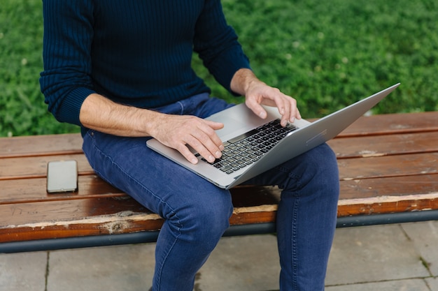 Close up of male freelancer sitting bench and working on laptop. Young man in casual clothing typing on portable computer outdoors.