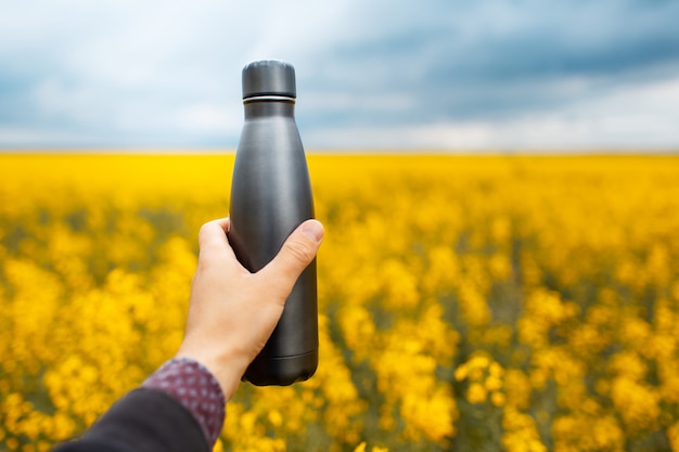 Close up of male hand holding a dark grey, eco metal bottle on background of blurred rapeseed field.