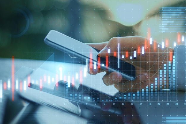 Close up of male hand using cellphone and tablet at desktop with abstract glowing candlestick business chart on blurry outdoor background Trade finance and market concept Double exposure