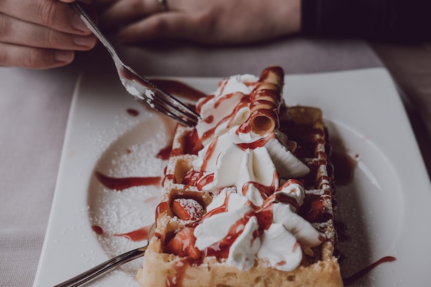 Close up di mani maschili che tengono una forchetta mangiando un waffle con crema fresca e salsa di fragole