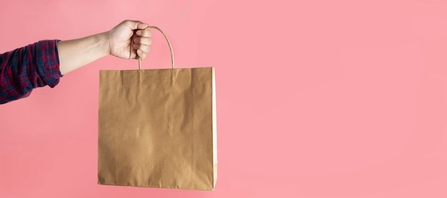 Close up male holding in hand brown clear empty blank craft paper bag for takeaway isolated on pastel pink background