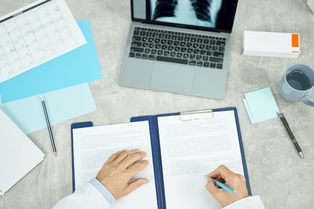 Крупным планом рук мужского врача, делающего документы, в то время как человек сидит за столом с блокнотом, бумагами и чашкой кофе