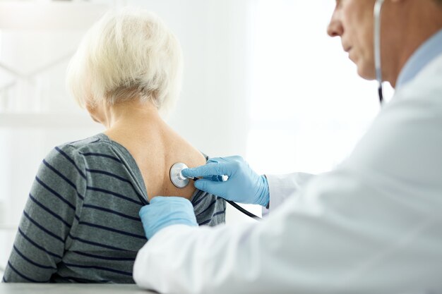 Close up of male physician in sterile gloves putting stethoscope on old woman back and checking her breath