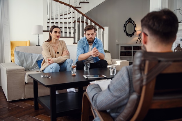Close up of male psychologist sits at chair and listens depressed young couple at stylish cozy office. Selective focus.