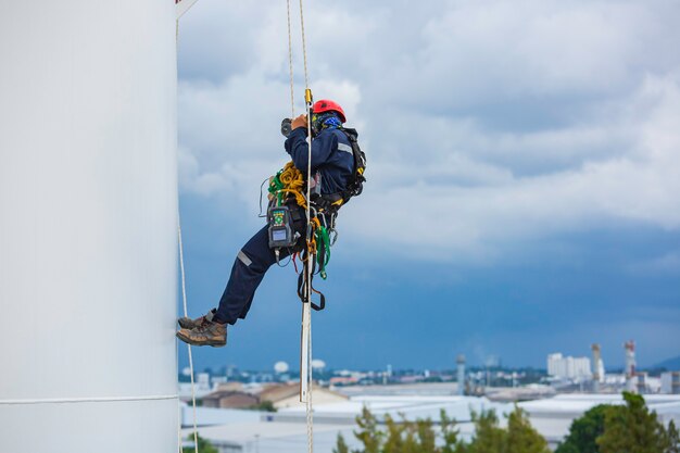 Close-up male workers control equipment rope down tank rope\
access inspection of thickness shell plate storage tank gas\
background cloud storm.