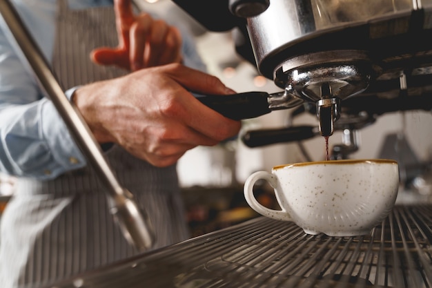 Close up of man bartender hand holding handle of coffee machine and pouring hot beverage into white cup from the tap