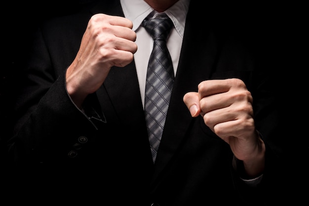 Close up of man in black suit with hand gesture on black background.