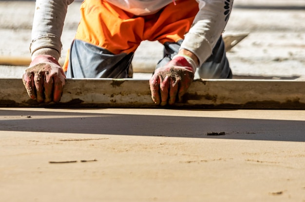 Close up of man builder placing screed rail on the floor covered with sand-cement mix at construction site. Male worker leveling surface with straight edge while screeding floor. Blurred background.