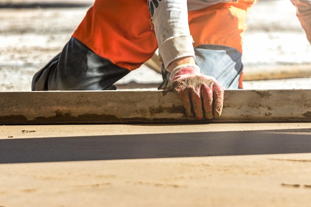 建設現場で砂セメント ミックスで覆われた床にスクリード レールを配置する男性ビルダーのクローズ アップ背景をスクリードしながらストレート エッジで表面を平準化する男性労働者