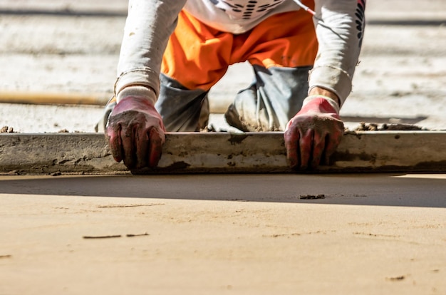 Close up of man builder placing screed rail on the floor covered with sandcement mix at construction site Male worker leveling surface with straight edge while screeding floor Blurred background