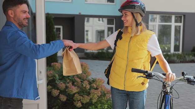 Close up man customer open door to a food delivery woman\
wearing yellow thermal backpack on a bike and recieves order or\
package with food food delivery concept