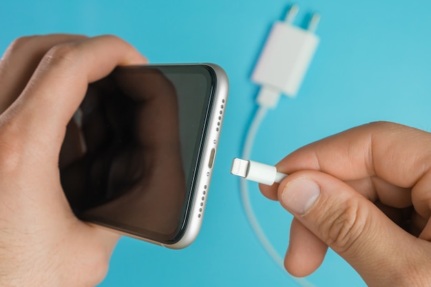 Close-up man hand invoegen van een oplader in de aansluiting van de mobiele telefoon via bliksem USB A-kabel op het blauwe oppervlak.