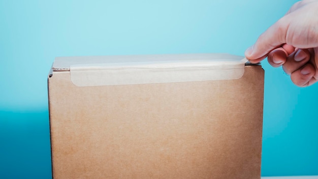 Photo close up man hand's opening cardboard box package by pulling tear tape unboxing brown cardboard box tearoff tape gift and delivery box box is lying on a white table with a blue background