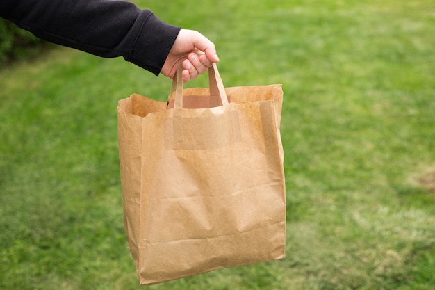 Close up of man hand with eco paper bag for takeaway food on nature green background. Delivery in any weather around the clock to the client.