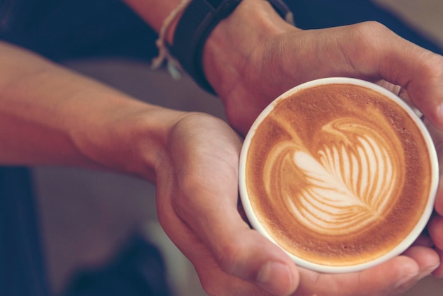 カフェのコーヒーショップでラテアートコーヒーカップを持っている男の手を閉じる男の手でホットコーヒーマグミルクラテアート新鮮な朝の時間にリラックス